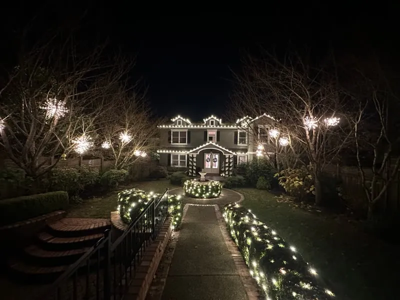Two-story home with warm white roofline lights, starburst lights in the trees and net-lit hedges along the front walk