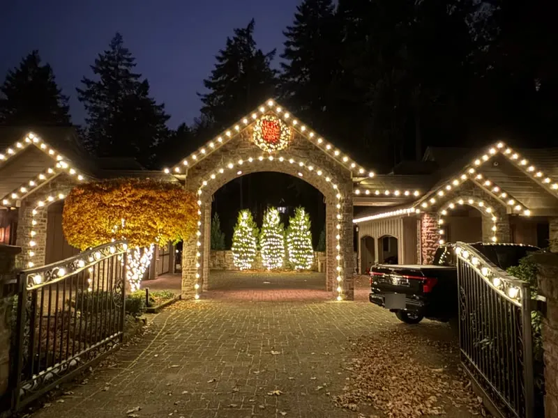 Warm white C9 lights outlining a stone archway and rooflines, with three light-wrapped evergreen trees beyond the arch